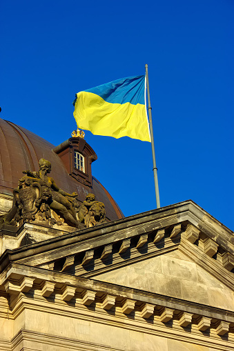 The Ukrainian flag flies against a blue sky atop a German museum building decorated with statues in Berlin, Germany