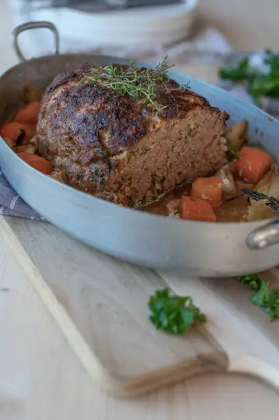 Delicious homemade german pork meatloaf with vegetables in a rustic roasting pan on pine wood table background with cross section view.