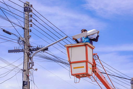 Electrician in bucket boom truck is repairing street light pole against blue sky background