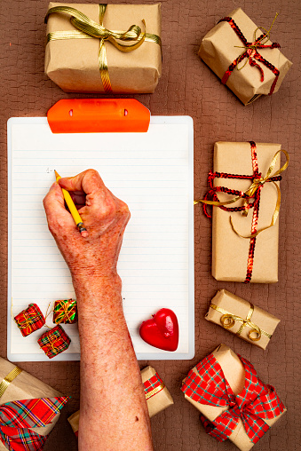 High angle view of white plastic clipboard for notes with Christmas gift boxes around it over brown fabric and mature man with pencil no make notes.