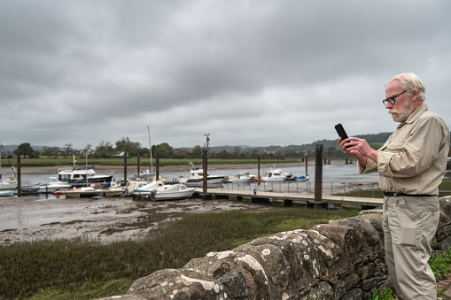 Retired man using a mobile phone while standing next to a wall at a marina in Scotland