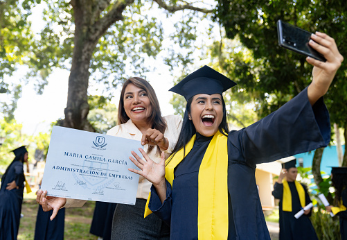 Happy Latin American woman taking a selfie with her mother holding her diploma on her graduation day - education concepts