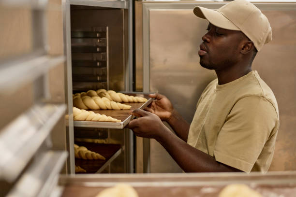 Side view portrait of Black young man putting tray in industrial oven while working in bakery, copy space