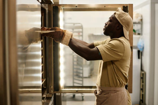 Black young man putting tray with fresh breads in oven at bakery kitchen