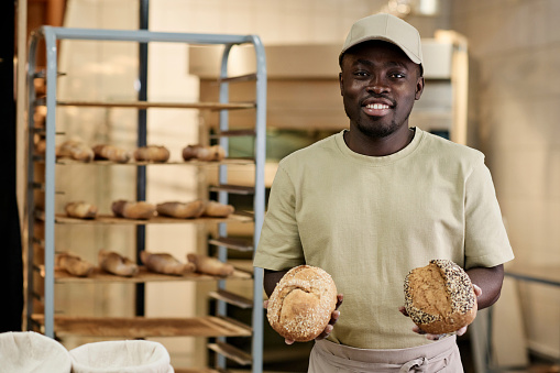 Waist up portrait of young Black man holding fresh whole wheat bread in bakery kitchen and smiling at camera, copy space