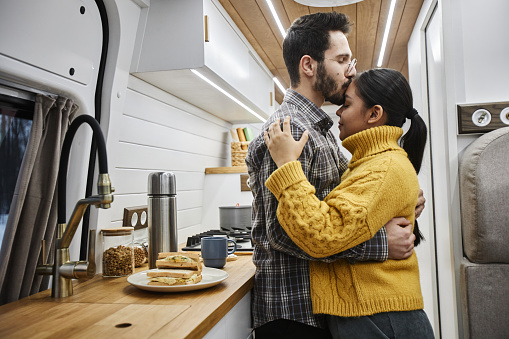 Side view portrait of young couple embracing affectionately with husband giving kiss on forehead while standing in trailer van and travelling together, copy space