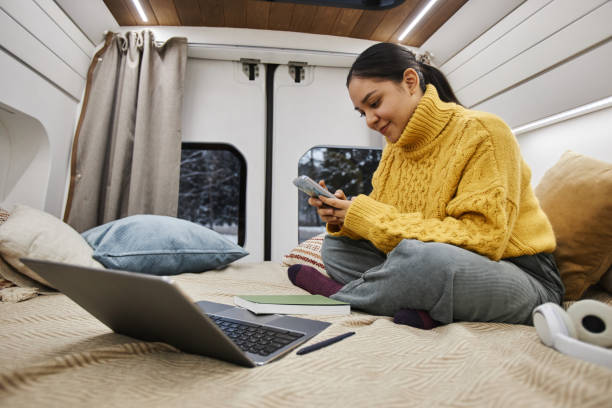 mujer joven con suéter acogedor usando un teléfono inteligente en la cama en una camioneta de remolque - mobile home audio fotografías e imágenes de stock