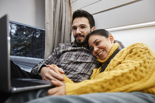 Portrait of smiling young couple in trailer van using computer on bed together while enjoying cozy travelling in winter