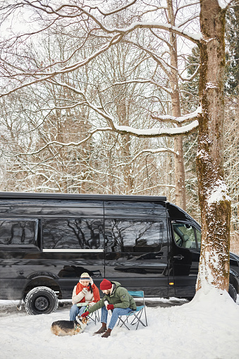 Vertical portrait of young couple travelling by van in winter forest and petting cute dog