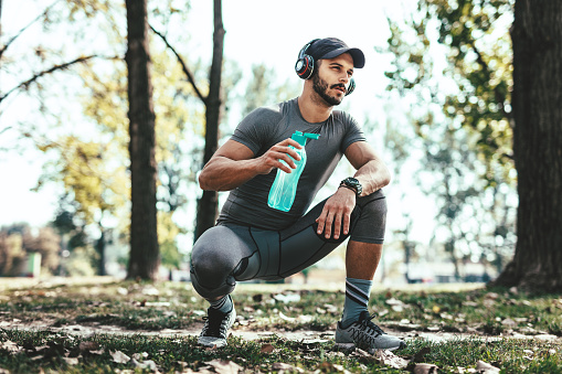 Young man with headphones is resting after training in autumn nature. He is holding a bottle of water and listening music.
