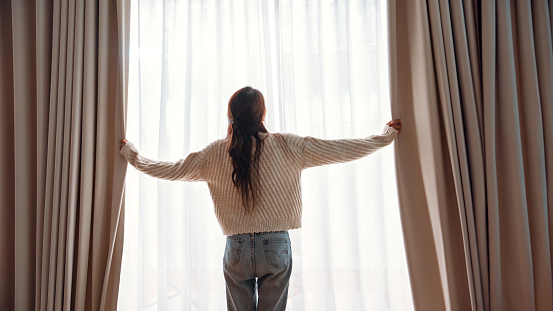 Back view - Asian woman opening curtains at morning, Hands pulling window curtain