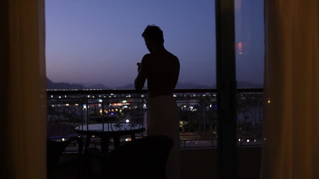silhouette of a man in the evening on a balcony overlooking the city and mountains uses a smartphone.