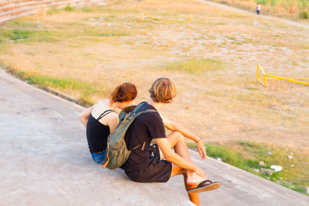 Young white backpacker couple is sitting  at Mekong river. They  are sitting and relaxing on promenade in Vientiane. They are wearing casual clothings and are relaxing at border of promenade. Behind and below  is wetland at river. They are relaxing and talking