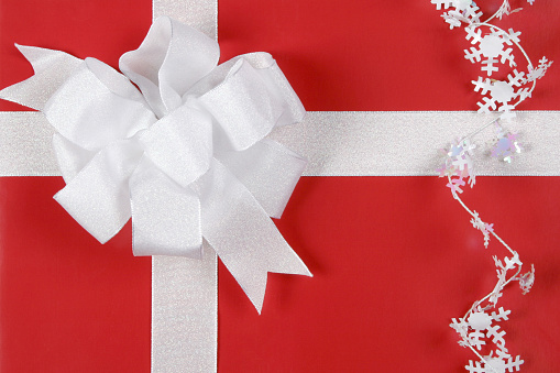 A beautiful red gift box tied with white ribbon and a white bow with snowflake confetti.