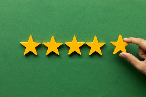 Five star rating feedback. Hand is holding  last star shape in front of green colored background. Giving best score point to review the service