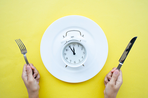 Directly above view of white plate with alarm clock and  person holding fork and knife  on yellow background. Representing diet, Intermittent fasting, time-restricted eating and healthy food concept.