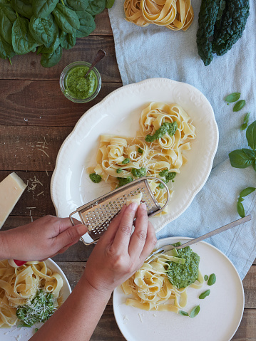 Hands grating Parmesan cheese over a platter of Tagliatelle pasta with spinach pesto and kale. Traditional Italian meal.