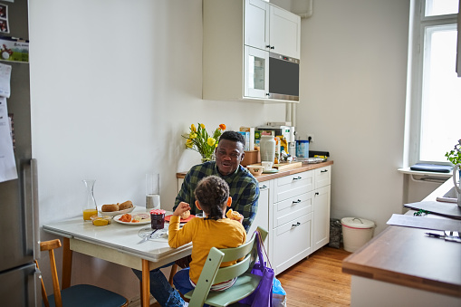 African man with son having breakfast sitting at table in kitchen. Father talking with son having breakfast at dining table in kitchen.