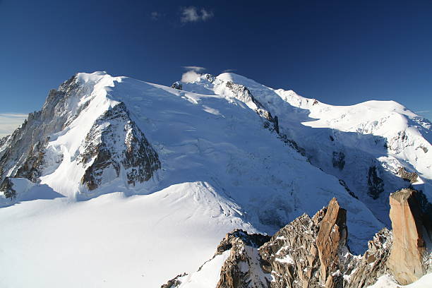 A striking view of Mont Blanc du Tacul reveals rugged rock faces emerging from pristine glacier ice under a vivid blue sky, with crisp sunlight accentuating the alpine textures.