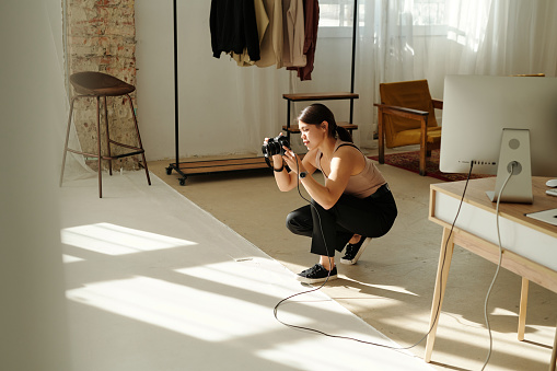 Young brunette woman with photocamera taking pictures of model while sitting on squats in the center of spacious studio during photo session