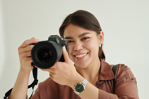 Happy young female photographer with photocamera taking pictures in isolation during photo session in studio and looking at you with smile