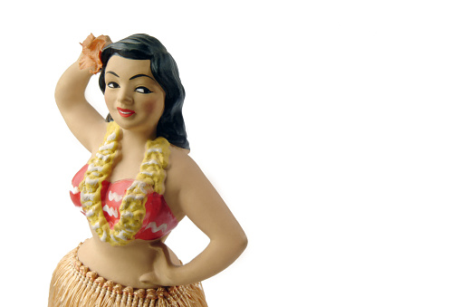 Close up image of antique Hawaiian hula girl figurine on white background. The figure is located in the left 1/3 of the image. The figure has black wavy hair, a red and white bikini top, and a yellow lei. The photo cuts off just below her hips so that only the top of her hula skirt is visible. She has one hand on her hip and one on her bed. 