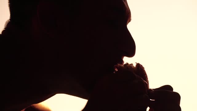 Silhouette of a man who eats a hamburger. Side view portrait of hungry person eating fatty burger with great appetite, fast food