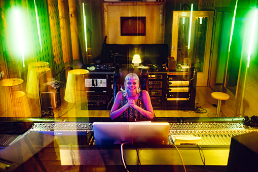 Portrait of a talented, experienced music producer, sitting in front of a mixing console in a music studio. Behind her are various pieces of equipment needed for recording and sound processing.