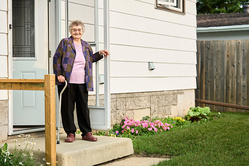 Portrait of a senior woman smiling while standing with a walking cane at the open front door of her house