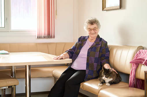 Smiling senior woman petting her cute tabby cat while sitting at her kitchen table at home