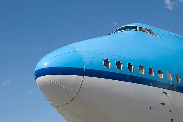 Nose and windows of a boeing 747 400 airplane, showing the cockpit windows on the upper deck and part of the main deck. The airplane is painted in blue and white colors and matches the clear blue sky.