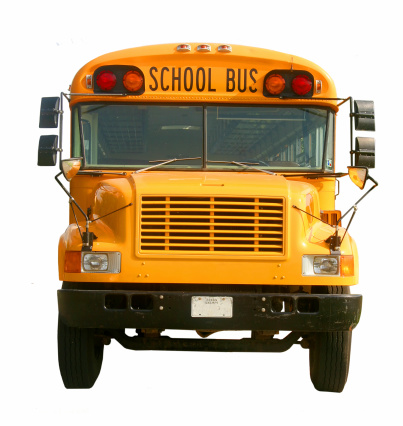 A close up of the front end of a traditional American yellow school bus outside an elementary school with large sign and safety lights in a city environment