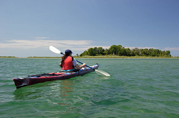A woman paddles her kayak towards a tree-lined coast on a clear, sunny day. Lake Michigan.