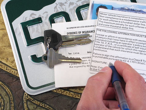 Using a Colorado license plate as a background, this color photo shows a driver’s papers related to proof of automobile insurance along with keys and a partial driver's license. The driver is signing the proof of insurance agreement on the back of the auto registration.