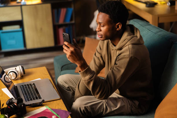 istock Concentrated serious African dark skinned black video production assistant, using his phone to communicate with colleagues and capture behind-the-scenes footage while editing raw footage on laptop 1726514419