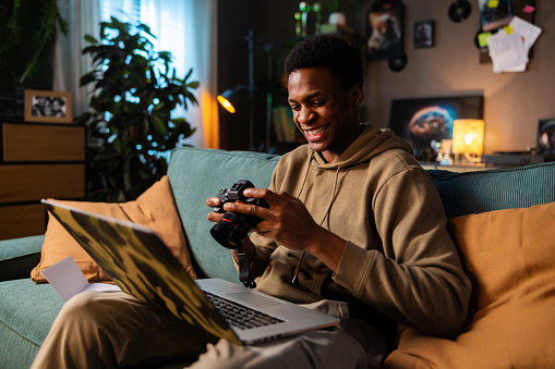 Carious delighted African American dark sinned black guy earing casual outfit sitting on sofa in cozy living room holding camera in hands observing photos while working online in laptop.
