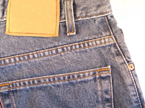 This color photo shows a closeup view of the back pocket of dark-blue, denim blue jeans. Above the pocket, a leather-like blank jeans label with red stitching allows you to add your own text.
