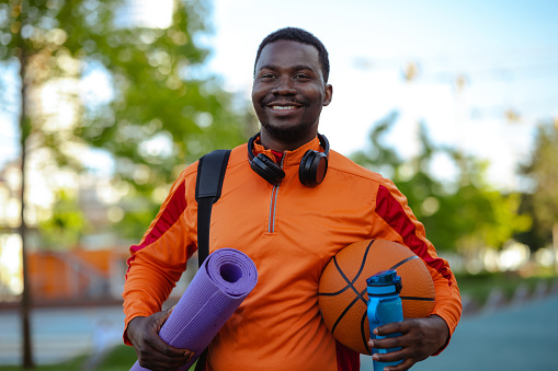 A young and cheerful African American man carrying sports equipment with a big smile on his face. He has wireless headphones around his neck.