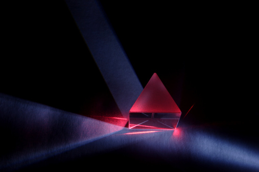 A triangular glass prism refracts a blue light and a red laser beam. The prism has a frosted top and sits on a lightly textured surface.