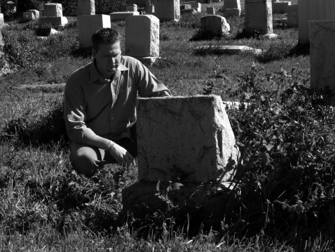 A man kneels beside a grave in an old cemetery. He looks forlorn.