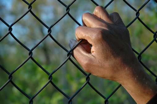 A male African-American hand clings to a chain link fence. Background out of focus.