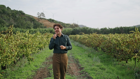 Happy relaxed farmer inspecting grape plantation walking on rural road between grapevine rows. Curly italian worker smiling enjoying beautiful landscape vineyard. Young winegrower satisfied harvesting