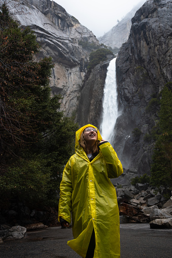 Smiling female hiker in a yellow raincoat standing in front of the amazing natural beauty of Ribbon Fall in Yosemite Valley