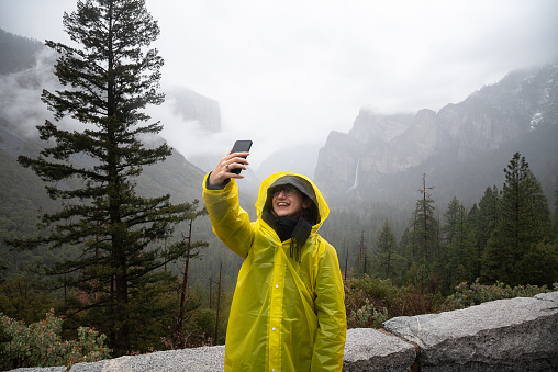 Smiling woman in a yellow raincoat taking a selfie from the iconic Tunnel View, while behind her stretching the view of Yosemite Valley under the fog