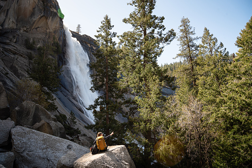 Woman hiker with a yellow backpack on her back siting on a rock under a tall fir tree and Nevada Fall