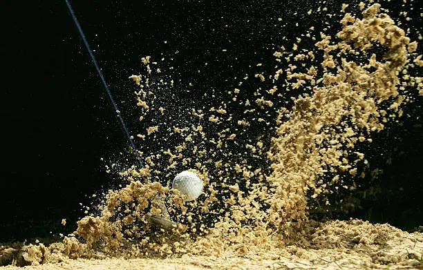 Sand flies up as a golf ball in a bunker is struck by a wedge in  front of a black background