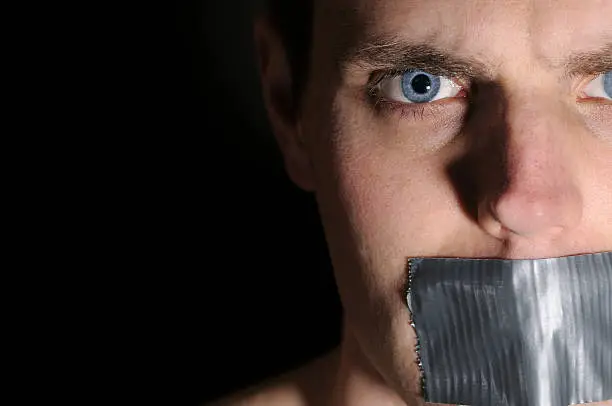 "A young man with duct tape over his mouth on a dark brown background. Studio shot, dramatic lighting. Copyspace."