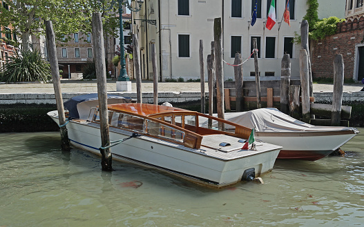 Two water taxis moored along the Grand Canal, Venice, Italy.   One covered.   White with brown top.   Large, plain poles.   Brown water.