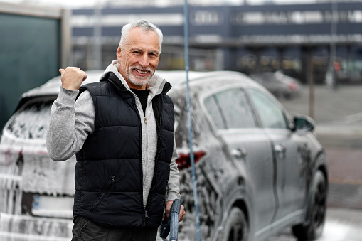 Smiling Caucasian stylish senior gray-haired man driver smiling a toothy smile at camera, showing his car in detergent foam, being washed in the car wash self service. Concept of keeping the car clean