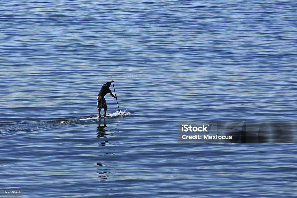 Surfboard Paddling A man stands on his surfboard and paddles across the open water. Beach Stock Photo